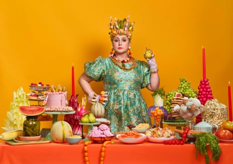 Siobhan McSweeney, in an ornate green dress and vegetable crown, stands amid a lavish food spread