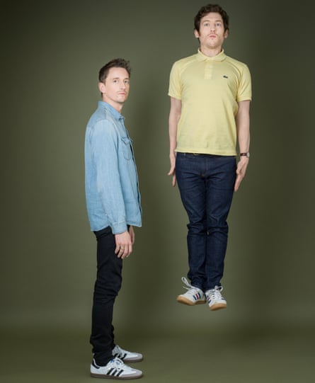 John Robins, standing side on, wearing a blue denim shirt and Elis James, jumping straight up, arms by his side, straight-faced, in a yellow polo shirt