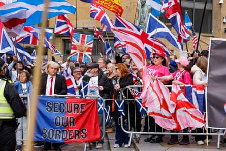 A group of people behind barriers wave union flags and the saltire. One person is dressed as Donal Trump holding a banner reading ‘secure our borders’