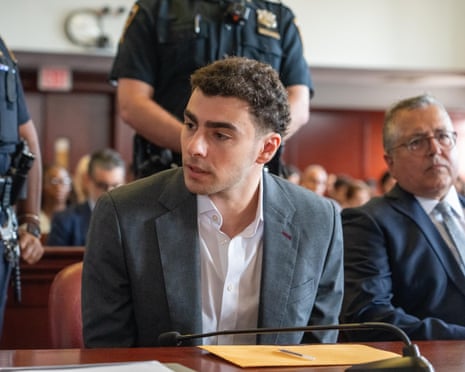 man in a grey suit sits at a courtroom table beside a police officer