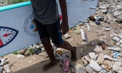 A child carries a surfboard, the ground is strewn with rubbish