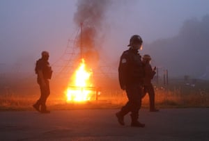 Police officers in riot gear walk past a bench on fire