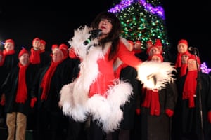 A woman dressed in a Santa-themed dress sings into a microphone with a group of background singers wearing matching hats and scarves