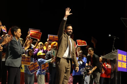 A man in a brown suit and tie smiles and waves on stage as a crowd cheers and claps behind him
