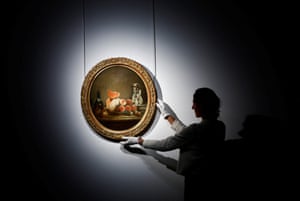 A woman wearing white gloves adjusts the hanging of  an oval-shaped still life of fruit