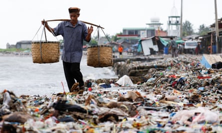 An elderly man carrying baskets walks through rubbish on a beach in Teluk fishing village, Indonesia