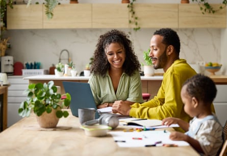 A man and a woman look happy as they sit at the kitchen table working on a laptop with their young child sitting nearby.