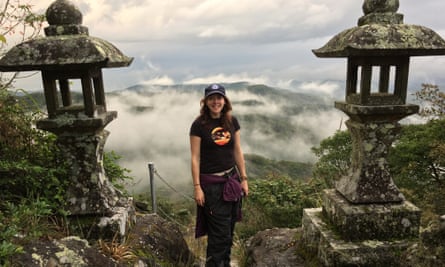 Emma John smiling, standing at a high point on her walk, two stone pillars either side of her, cloud cover below her in the background