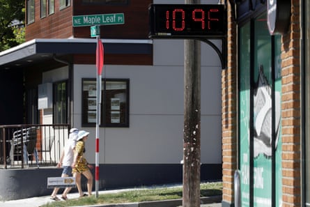 A digital sign on a shopfront in a Seattle street shows a temperature of 109F; two people wearing sunhats and shorts walk on the pavement in front of a white building in bright sunlight.