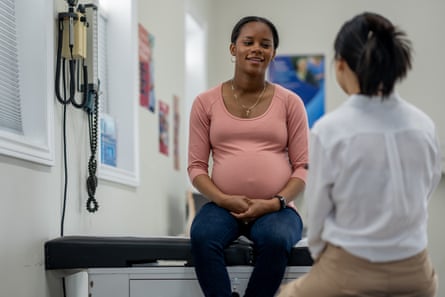 A pregnant woman is seated while conversing with a medical professional during a checkup.