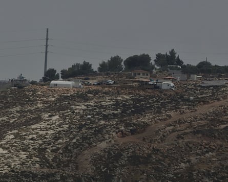 A rocky hillside in the West Bank with several small structures, vehicles and caravans scattered along the ridge, surrounded by sparse trees under an overcast sky. Dirt tracks wind across the foreground, and a utility pole stands to the left.
