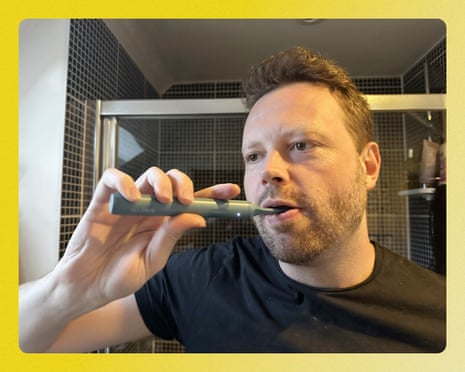 A man brushes his teeth with an electric toothbrush in a tiled bathroom.