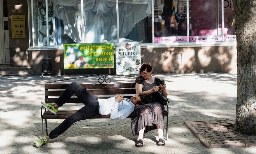 A young man in yellow trainers lies on a bench under a tree with his head in an older woman's lap, as she scrolls through her phone.