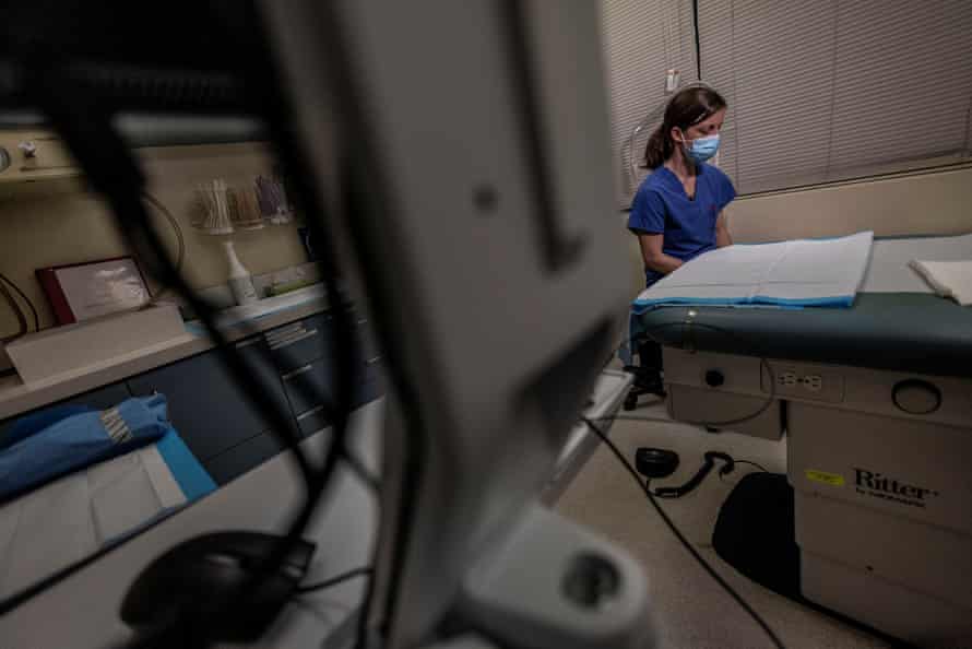 A woman in blue scrubs and a medical mask sits in an exam room. 