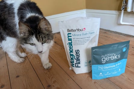 Iggy the cat walks next to a bag of Magnesium Bath Flakes and a bag of magnesium tables on a wooden floor