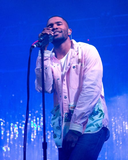 He holds a stand mic on stage and smiles against a sparking background