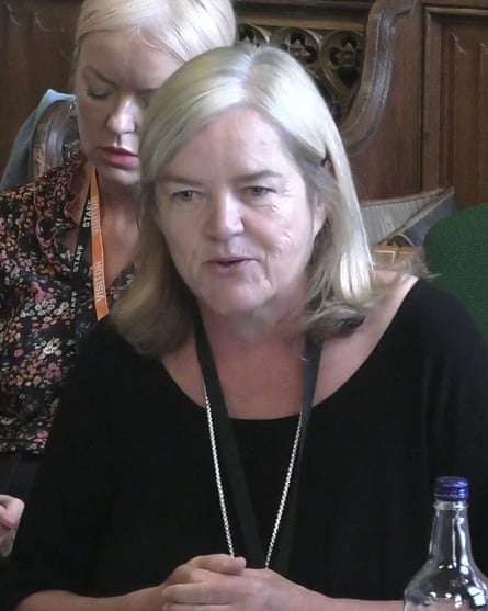 she has straight, shoulder-length blond hair and is about 60; she wears a black top and lanyard around her neck.