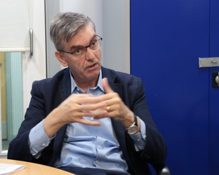 Thomas Keaney sits at a desk and raises his hands in front of him as he speaks. He has short grey hair and glasses, and wears a dark jacket and pale blue open-necked shirt.