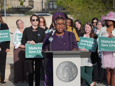 a woman dressed in purple at a lectern at a press conference with women standing behind her holding signs that read “midwives save lives”