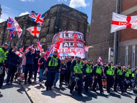 Police officers in hi-vis vests march in front of people holding union jacks and England flags with a big banner reading “Leave our kids alone” behind them