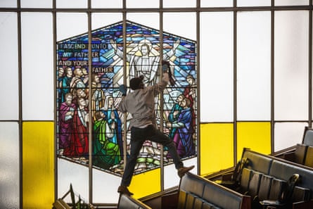 A man balances on the tops of church pews to polish a stained-glass wind