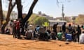 A group of people gather on an area of red dirt next to some trees and a wall, looking over an urban landscape