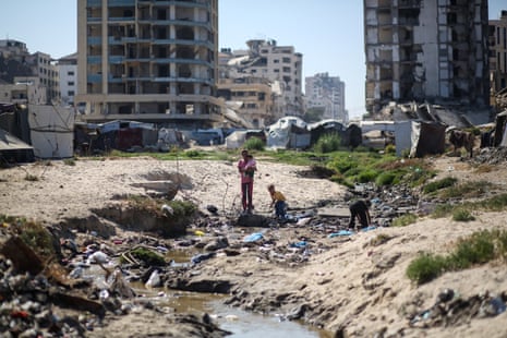 A woman and three children walking through a rubbish-strewn area, with bombed out buildings in the background.