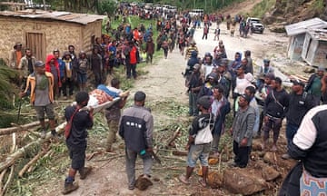 People carrying a person on a stretcher through a village after a landslide as many other people look on