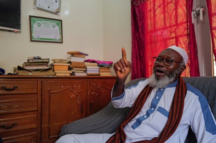A man in a white robe and hat speaks animatedly from an armchair.
