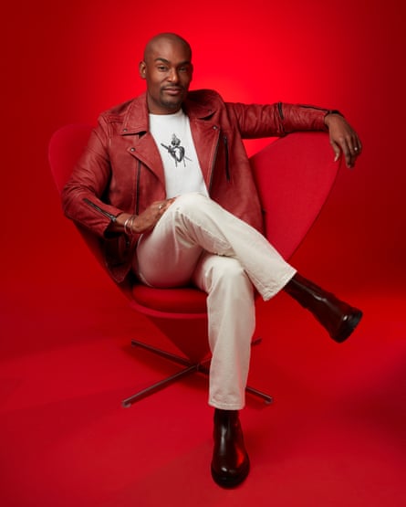 Paul C Brunson on a red background, sitting and leaning back in a red chair, wearing a white T-shirt, a cropped red leather jacket, cream jeans and black boots