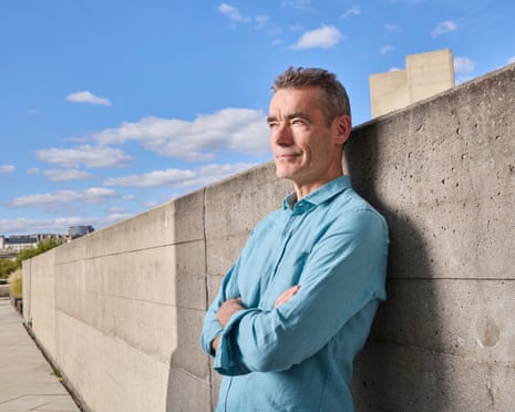 Rufus Norris leans against a concrete wall with his arms crossed, looking into the distance