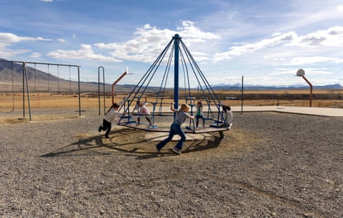 A wide shot of an old-fashioned merry-go-round, with cords attached from a center pole to the round edge, and five or six young people pushing it quickly, on a rocky flat yard with swings and a basketball hoop, under a huge blue sky, with low mountains beyond.