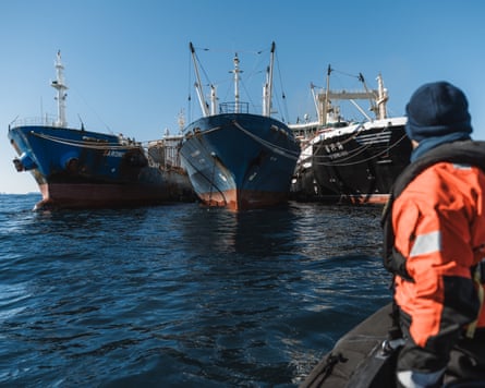 Three large ships on water. A man facing away from the camera in orange stands at the right-hand side of the image looking at the boats