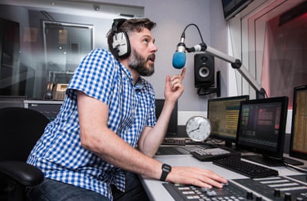Iain Lee in blue check short sleeve shirt and beard leaning into and talking into a mic in the studio at TalkRadio in 2016