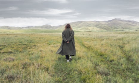 In a field a woman in an overcoat walks away from the camera towards mountains.