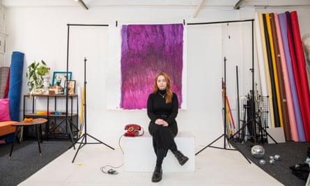 Camille Hewitt, in a long dress and thick-soled shoes, sits with her legs and hands crossed in front of an abstract painting hung from a pole in her art studio