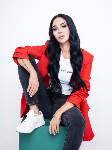 A woman in a white single top, with the words ‘Raise Your Standard’ on the front, and a red blazer, white trainers, and the words ‘female boss’ tattoed on her arm