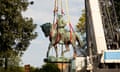 A greenish, oxidized statue of a man on a horse, wrapped and being hoisted by a crane under a hazy sky and amid trees.