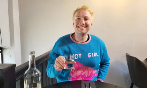 Joe Lycett holding a glass sitting at a restaurant table