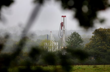 A tall white drilling rig seen through tree leaves in the green countryside