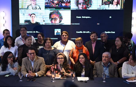 Two rows of people behind a desk with a microphone. Behind them is a screen showing other virtual attendees.