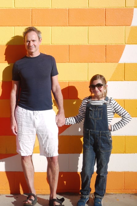 Paul Laity in white shorts and a blue T-shirt, and his daughter Martha, in dungarees and sunglasses, holding hands, with a yellow and orange brick wall behind them