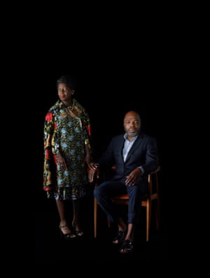 Portrait of Thelma Golden, who is standing dressed in  a beautiful dress of black and coloured prints of roses, beside Duro Olowu, who is sitting in a chair wearing a navy suit