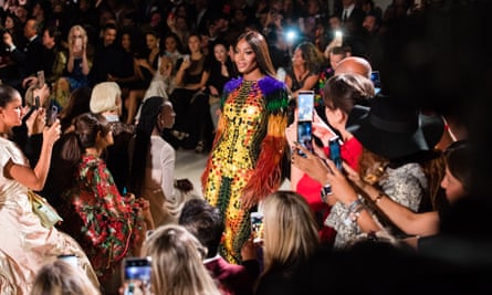 Naomi Campbell on the runway: she wears a glittering gold dress with large circular metallic pattern and purple, green, black and orange fluffy sleeves. She is surrounded by people taking photographs and filming her