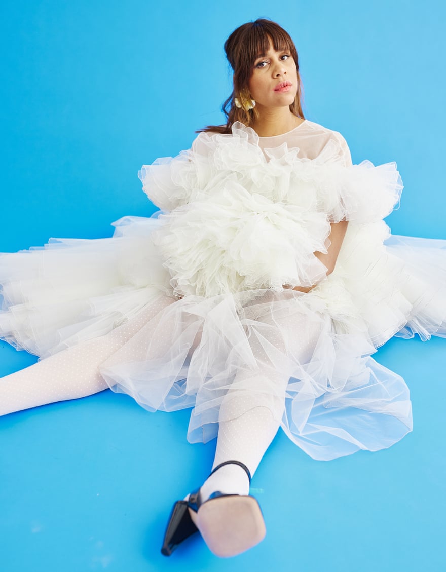Zawe Ashton sitting on the floor wearing a white dress with a very full skirt made up of layers of net