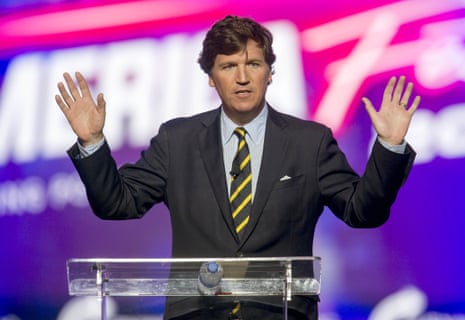 Tucker Carlson stands at a Plexiglass podium with both hands raised.