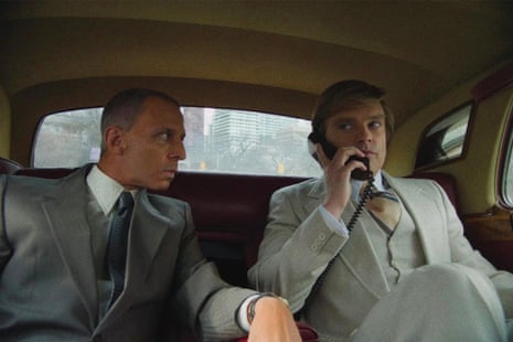 Two white men dressed in 80s-style suits sit in the back of a nice car.