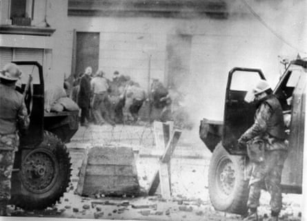 Black and white image of soldiers in helmets with guns standing behind the doors of sandbagged armoured cars while people run away towards a stone building; there are barbed wire and defences in front of the vehicles.