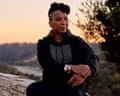 Kimberlé Crenshaw in a black hoodie sits on a stone wall at sunset with hills and trees behind her.