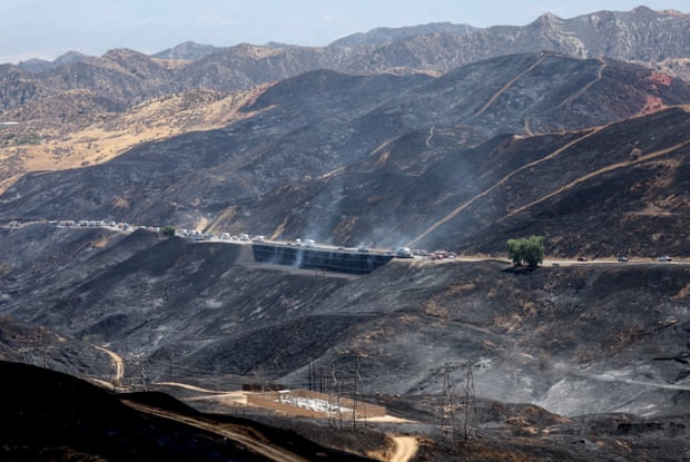 Vehicles traverse a highway through burned and smoking hills.